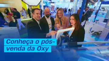 Equipe da Oxy conversando com visitantes em um estande, em volta de uma tela, com a chamada Conheça o pós-venda da Oxy